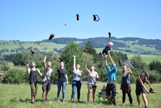 Kommunard*innen stehen auf einer Wiese und werfen Mützen und andere Dinge in die Höhe. Im Hintergrund Hügel, Wald und der Schafhof.